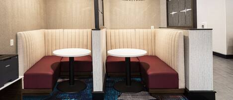 A modern hotel room with a corner seating area featuring two round tables and red cushioned benches.