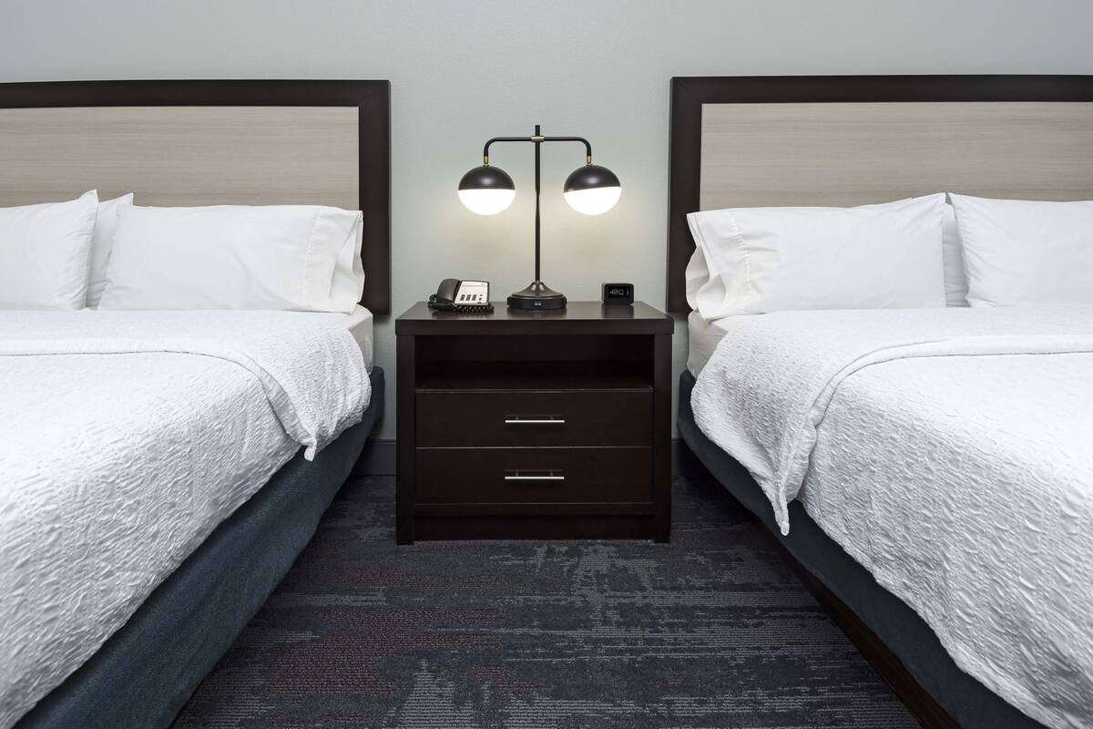 A hotel room with two beds, a nightstand with a lamp, a phone, and a clock.