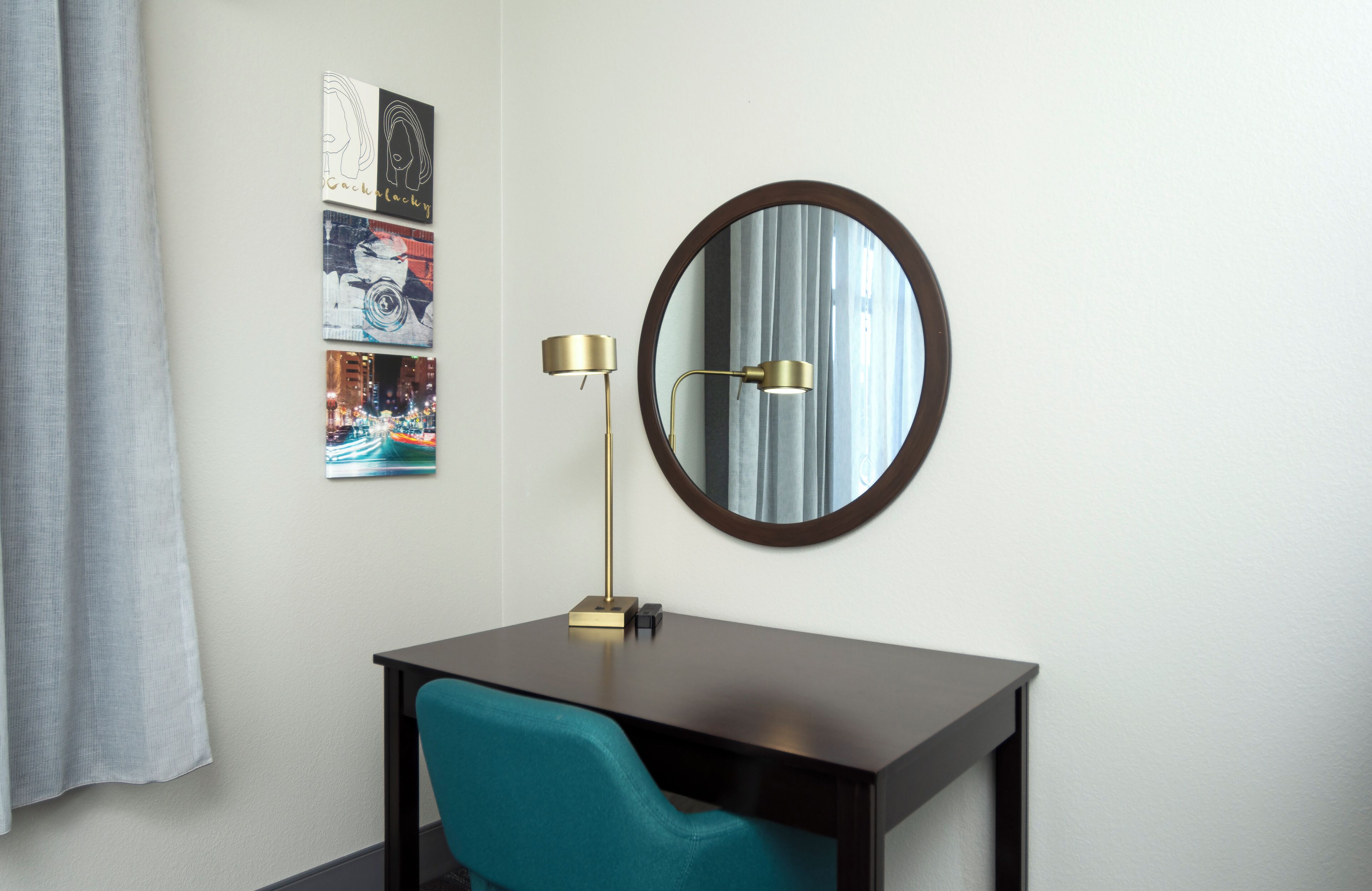 Une chambre d’hôtel équipée d’un bureau, d’une chaise de couleur turquoise, d’un miroir rond et d’une lampe fixée au mur.