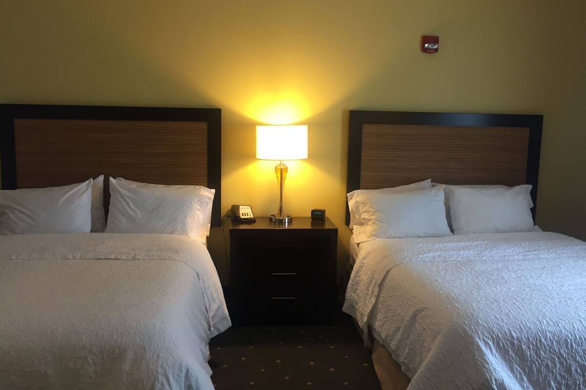 A hotel room with two beds, a bedside table with a lamp, a phone, and a fire alarm on the wall.