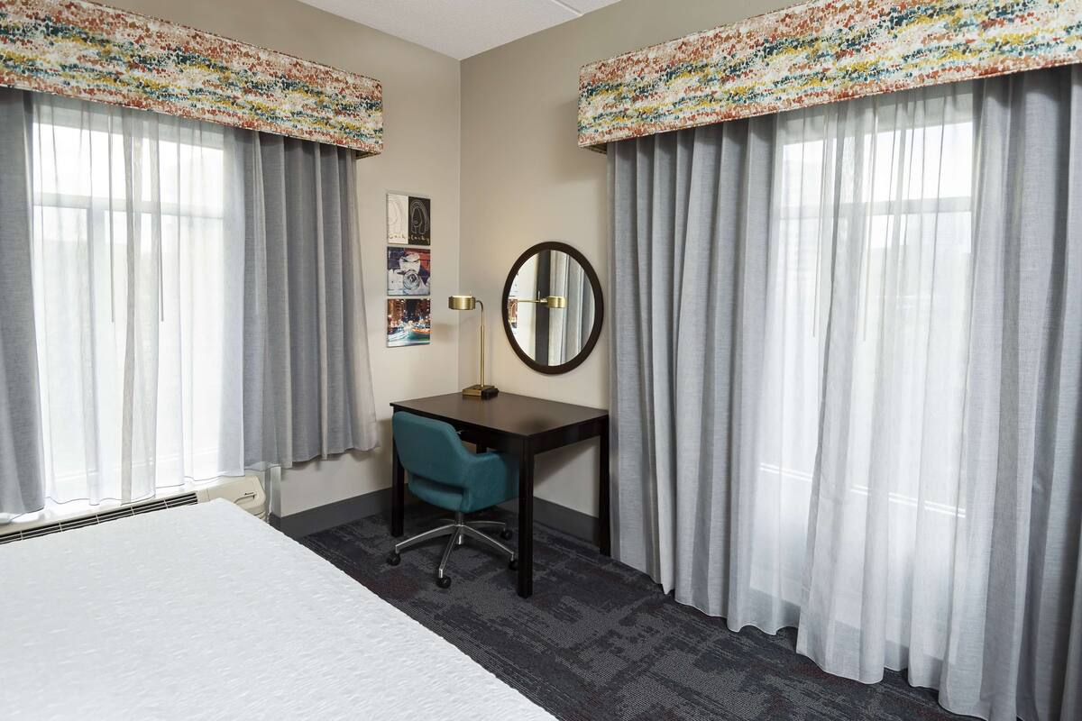 A hotel room with a bed, a desk with a chair, a round mirror, and two windows with curtains.