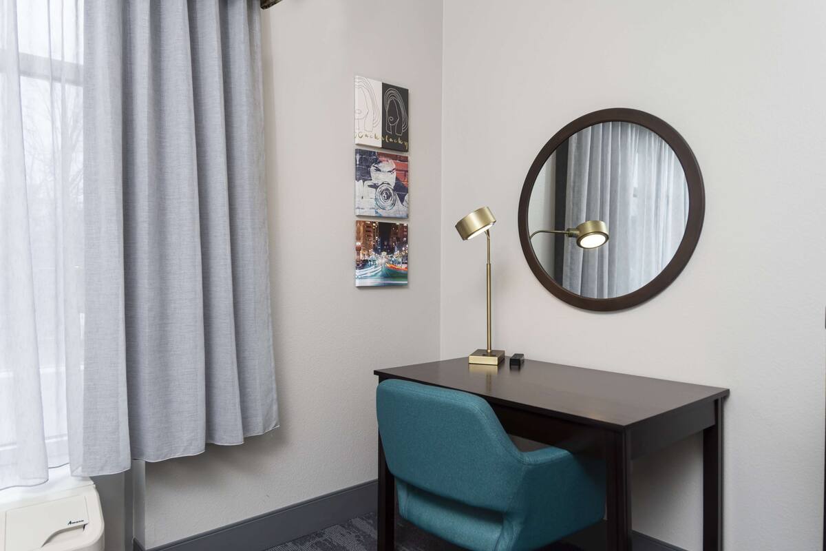 A hotel room with a desk, a teal chair, a round mirror, and three framed pictures on the wall.