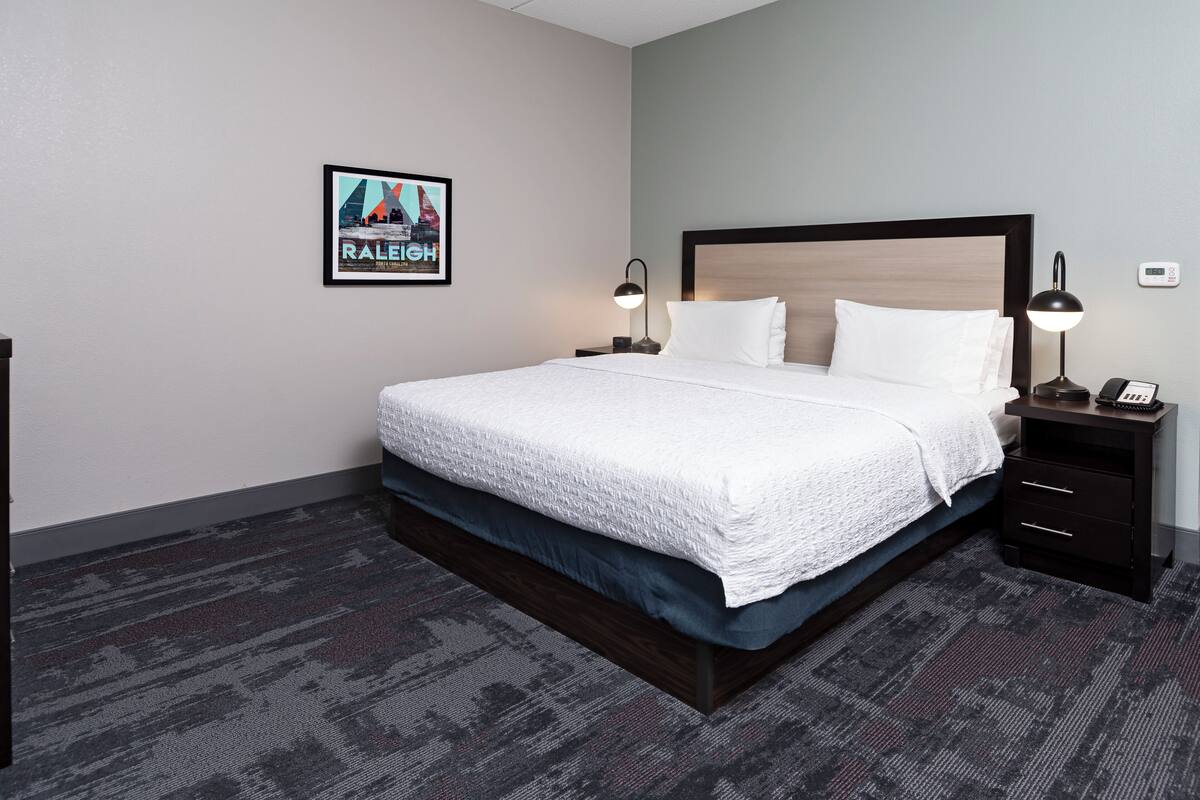 A hotel room with a large bed, two bedside tables, a framed picture on the wall, and a carpeted floor.