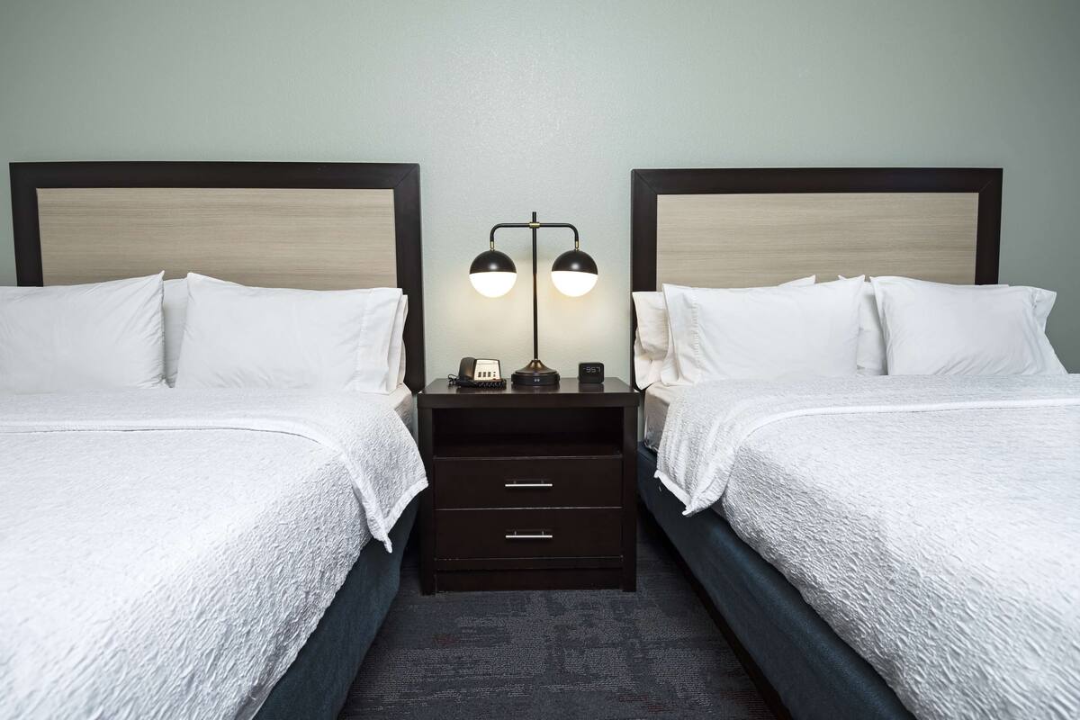 A hotel room with two beds, a nightstand with a lamp, and a small drawer unit.