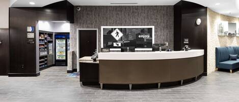 A modern hotel reception area with a curved reception desk, a digital screen displaying the hotel's logo, and a seating area with a blue sofa.