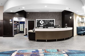 A modern hotel reception area with a curved reception desk, a digital screen displaying the hotel's logo, and a seating area with a blue sofa.