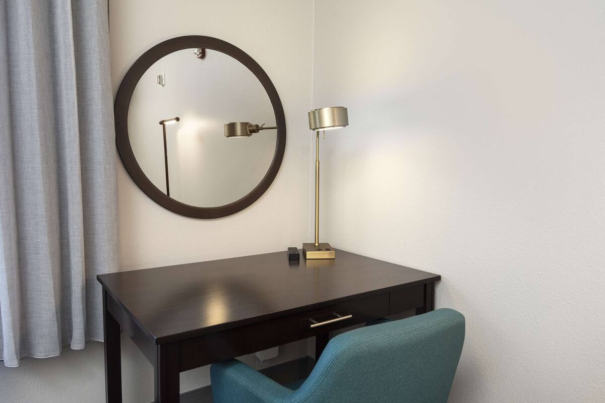 A hotel room with a dark wooden desk, a round mirror, a floor lamp, and a teal chair.