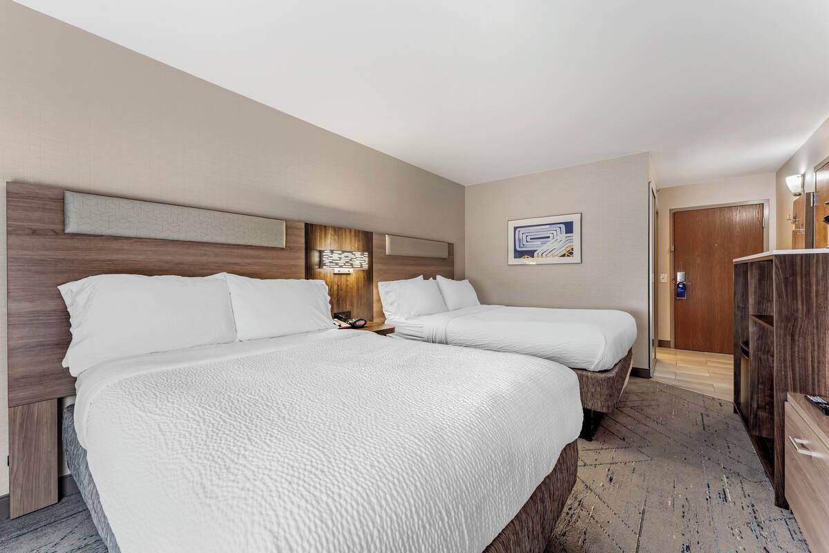 A hotel room with two beds, a wooden headboard, a nightstand, a wall-mounted light, and a door leading to another room.