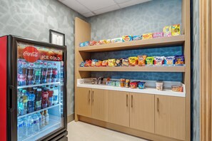 Máquina expendedora con un rótulo de Coca-Cola, estantes llenos de aperitivos y bebidas, y un mueble de madera con diferentes alimentos.