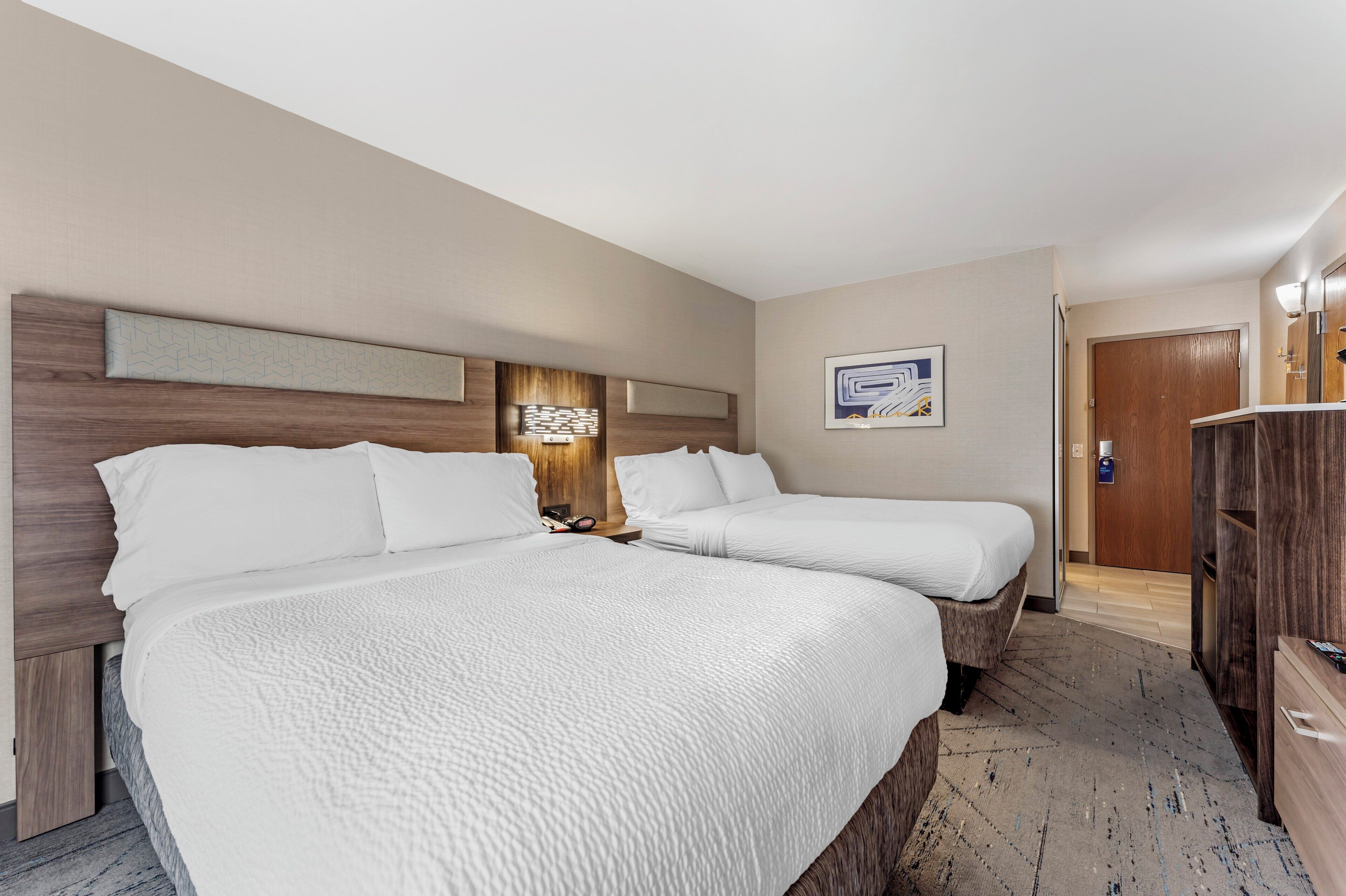 Habitación de hotel con dos camas, cabecera de madera, mesita de noche, lámpara de pared y puerta que da a otra habitación.