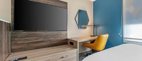 Una habitación de hotel moderna con una televisión de pantalla plana grande montada en la pared, un escritorio de madera con una silla, una cama con edredón blanco y una ventana con vistas.
