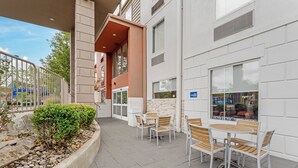 An outdoor patio area with a seating arrangement, a stone planter with greenery, and a building facade with windows and a door.