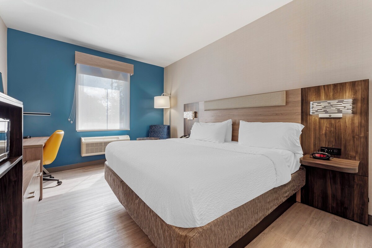 Una habitación de hotel moderna con una cama grande, un escritorio con silla, una mesita de noche con lámpara y un ventanal con cortinas.