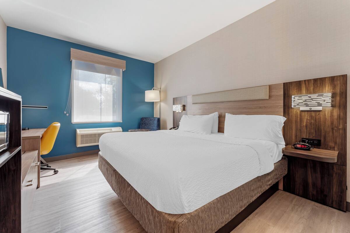 A modern hotel room with a large bed, a desk with a chair, a bedside table with a lamp, and a window with curtains.