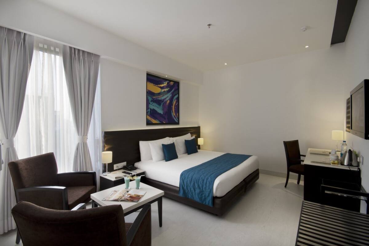 A hotel room with a bed, two chairs, a desk, a coffee maker, and a painting on the wall.