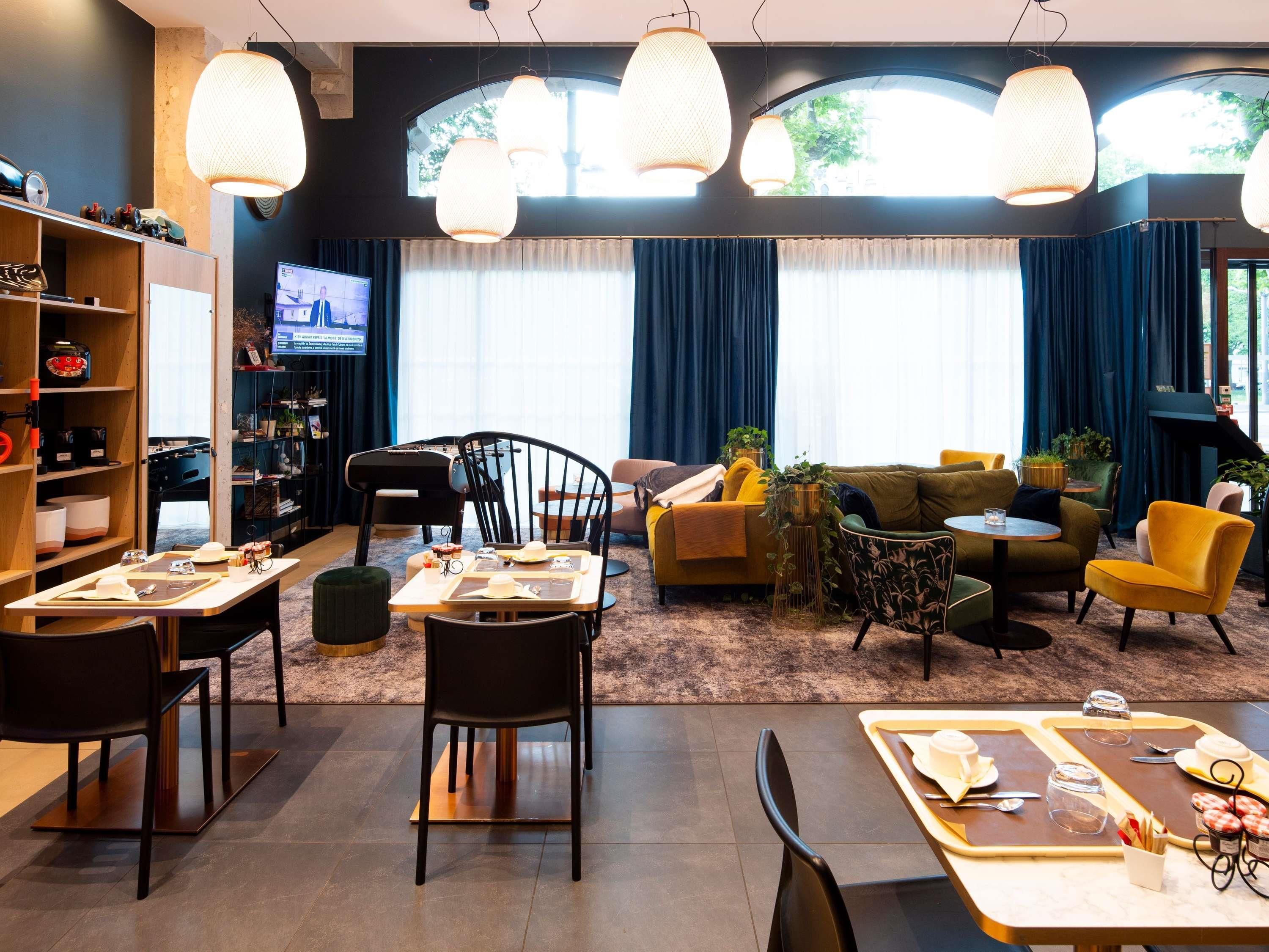 A modern hotel lobby with a dining area, sofas, and a TV screen.