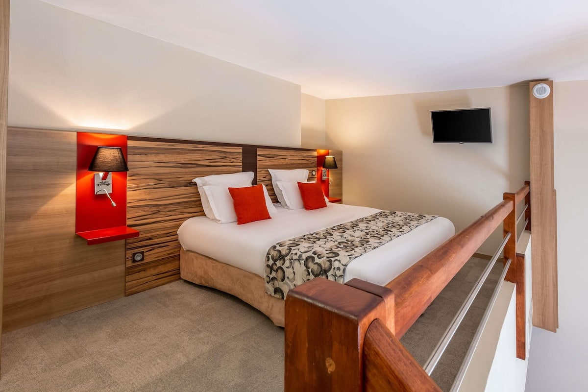 A hotel room with a bed, two bedside tables with lamps, a TV mounted on the wall, and a wooden headboard.