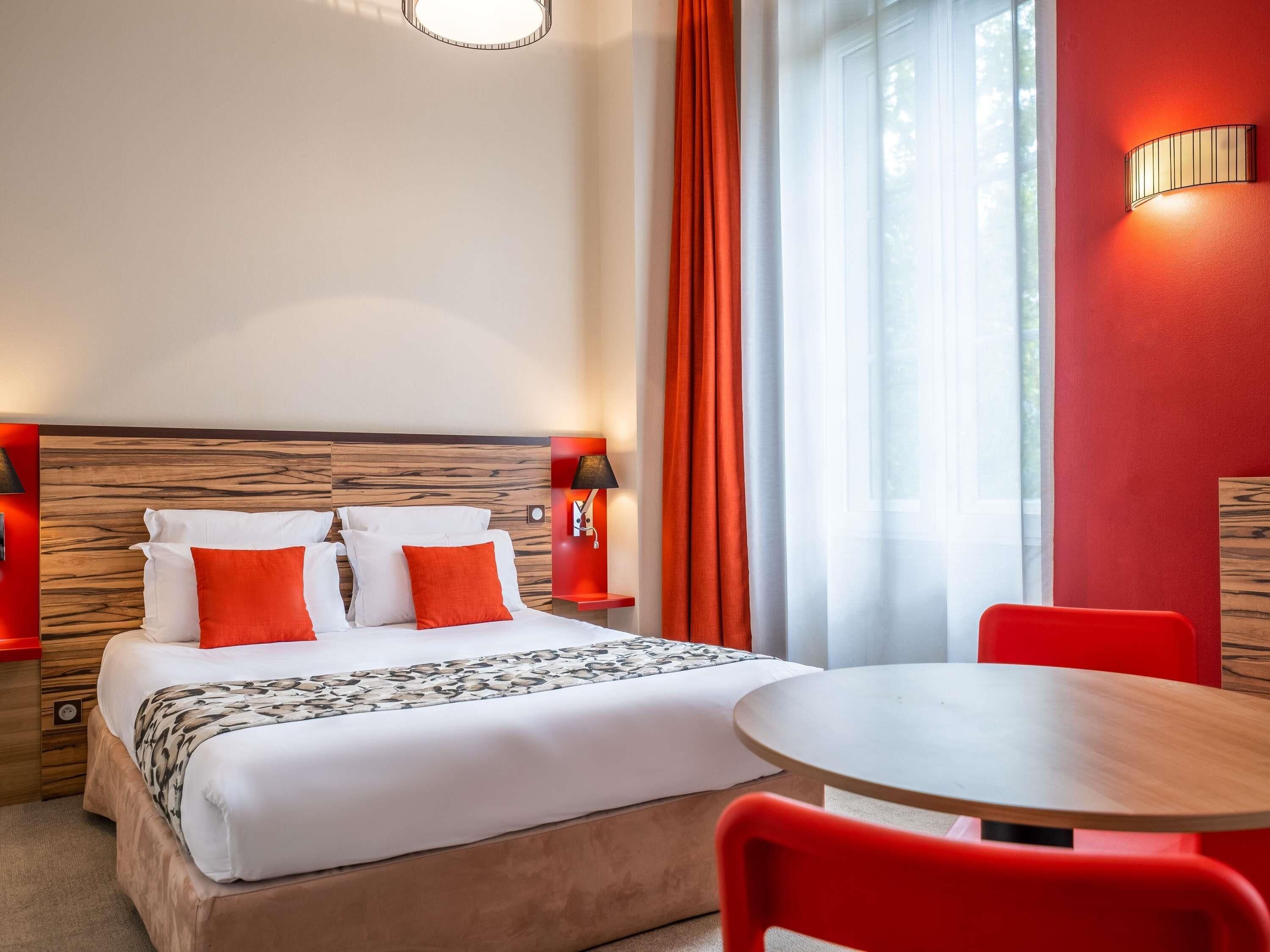 A hotel room with a bed, two bedside tables, a round table, and red chairs.
