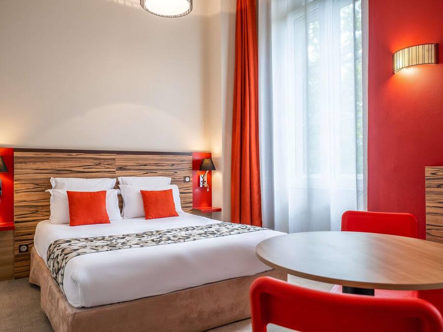 Une chambre d’hôtel avec un lit, deux tables de chevet, une table ronde et des chaises rouges.