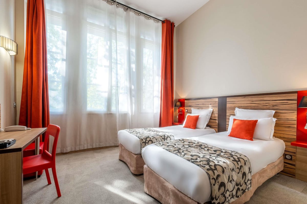 A hotel room with two beds, a desk, a chair, and a window with curtains.