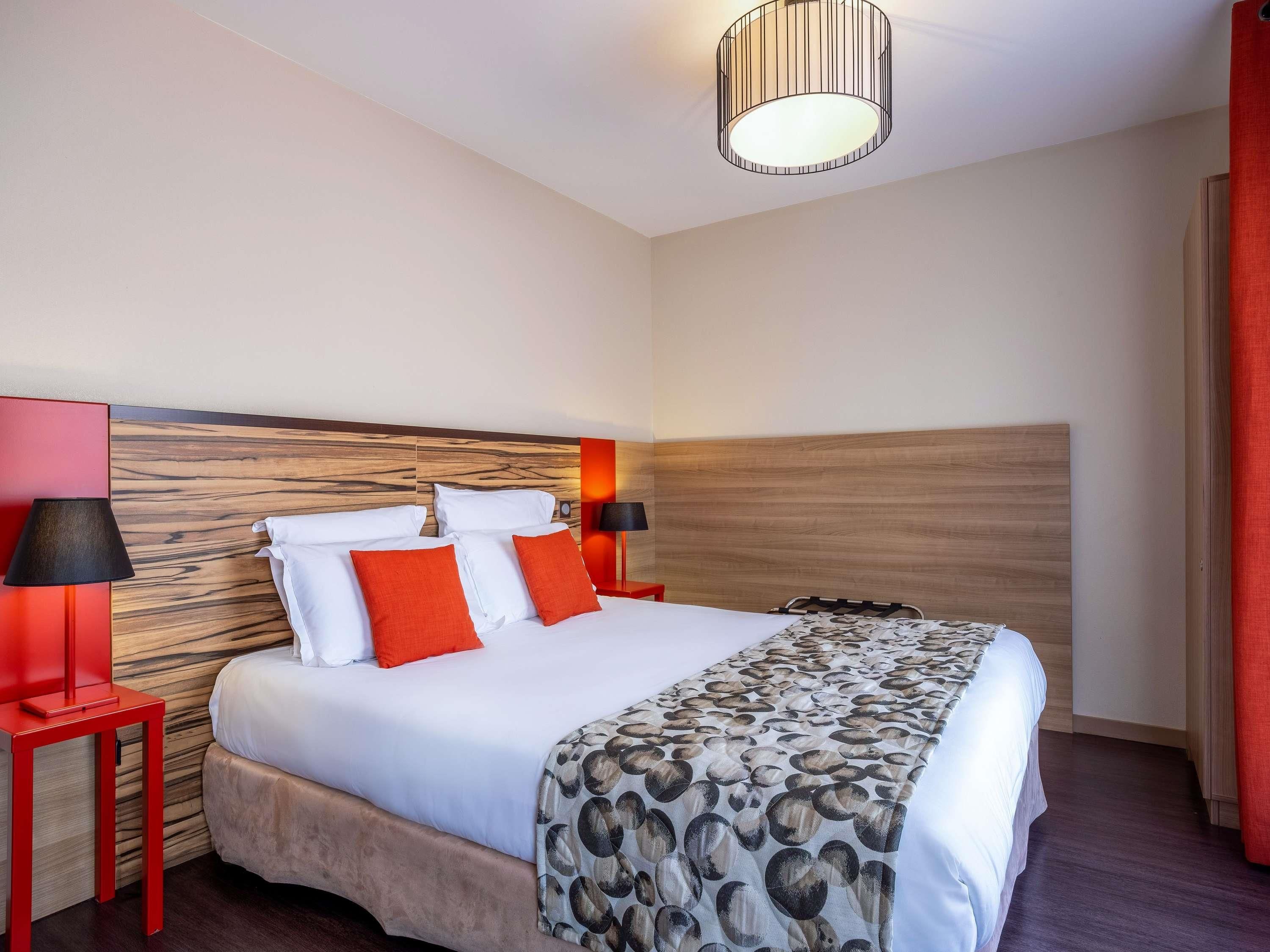 A hotel room with a wooden headboard, a bed with white and orange bedding, a patterned throw, and two bedside lamps.