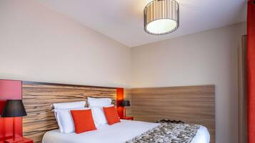 A hotel room with a wooden headboard, a bed with white and orange bedding, a patterned throw, and two bedside lamps.