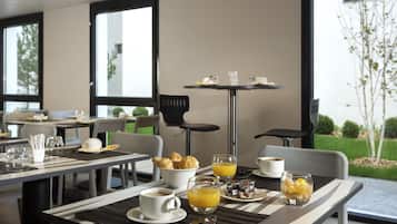 Uma mesa de pequeno-almoço com croissants, sumo de laranja, café e uma cesta de fruta, com vista para um jardim.
