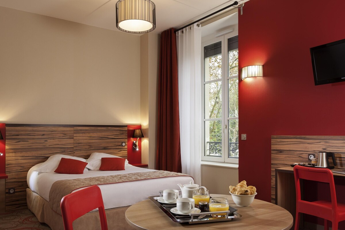A hotel room with a bed, a dining table with breakfast items, a TV, and a window with curtains.