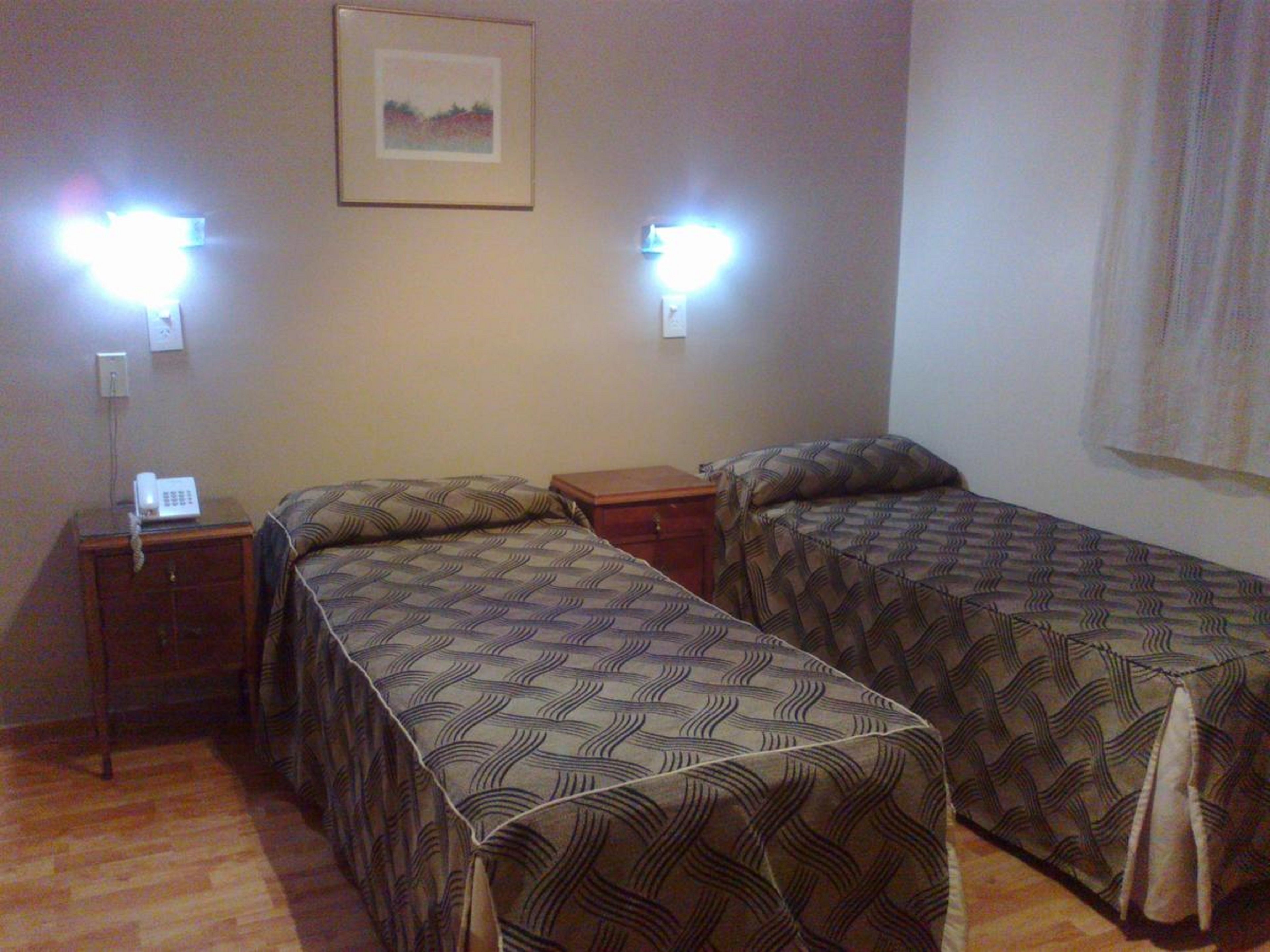 A hotel room with two beds, each with a patterned bedspread, a bedside table with a phone, and wall-mounted lights.