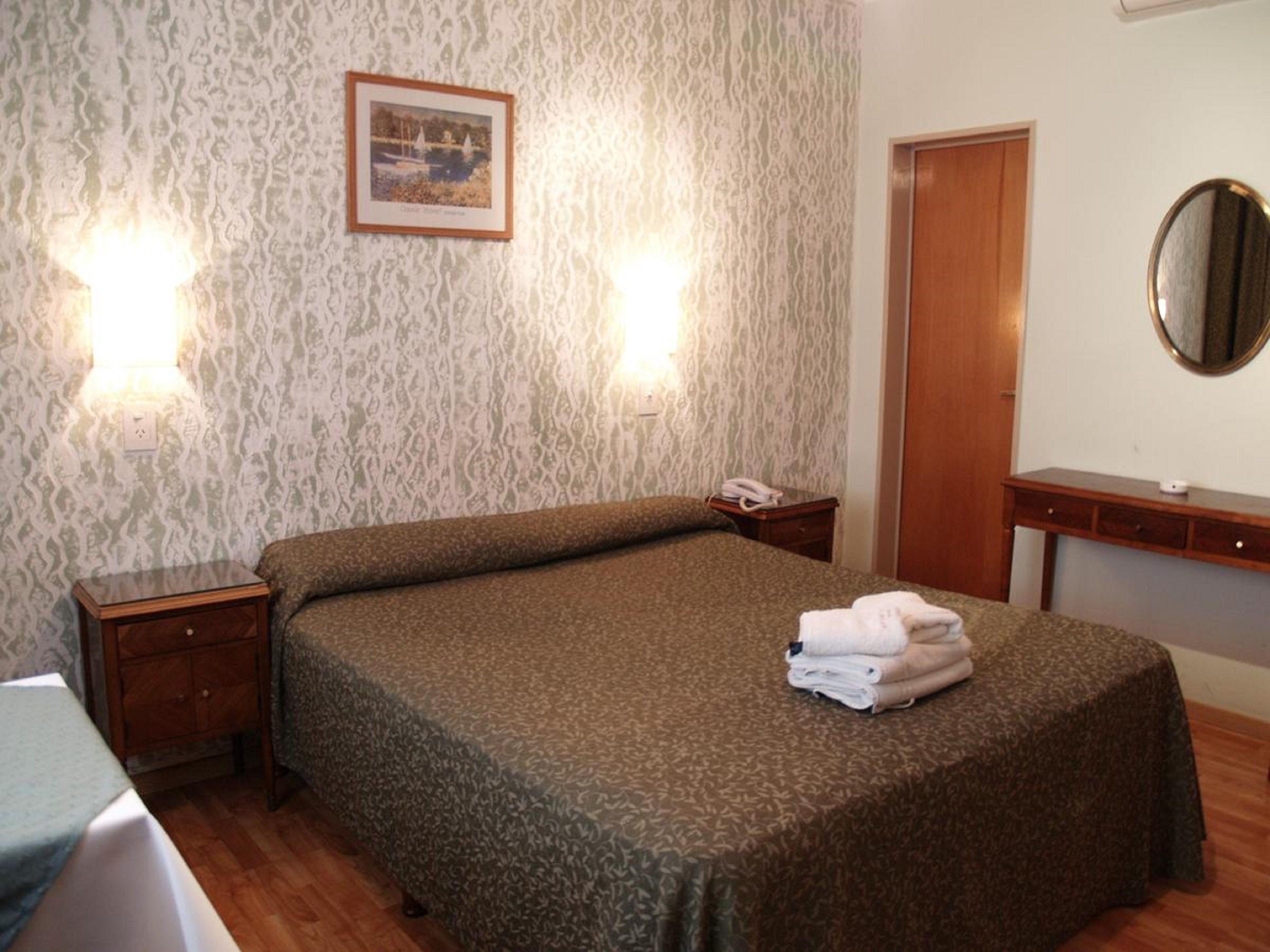 A hotel room with a patterned bedspread, a bedside table with a lamp, a mirror, and a framed picture on the wall.