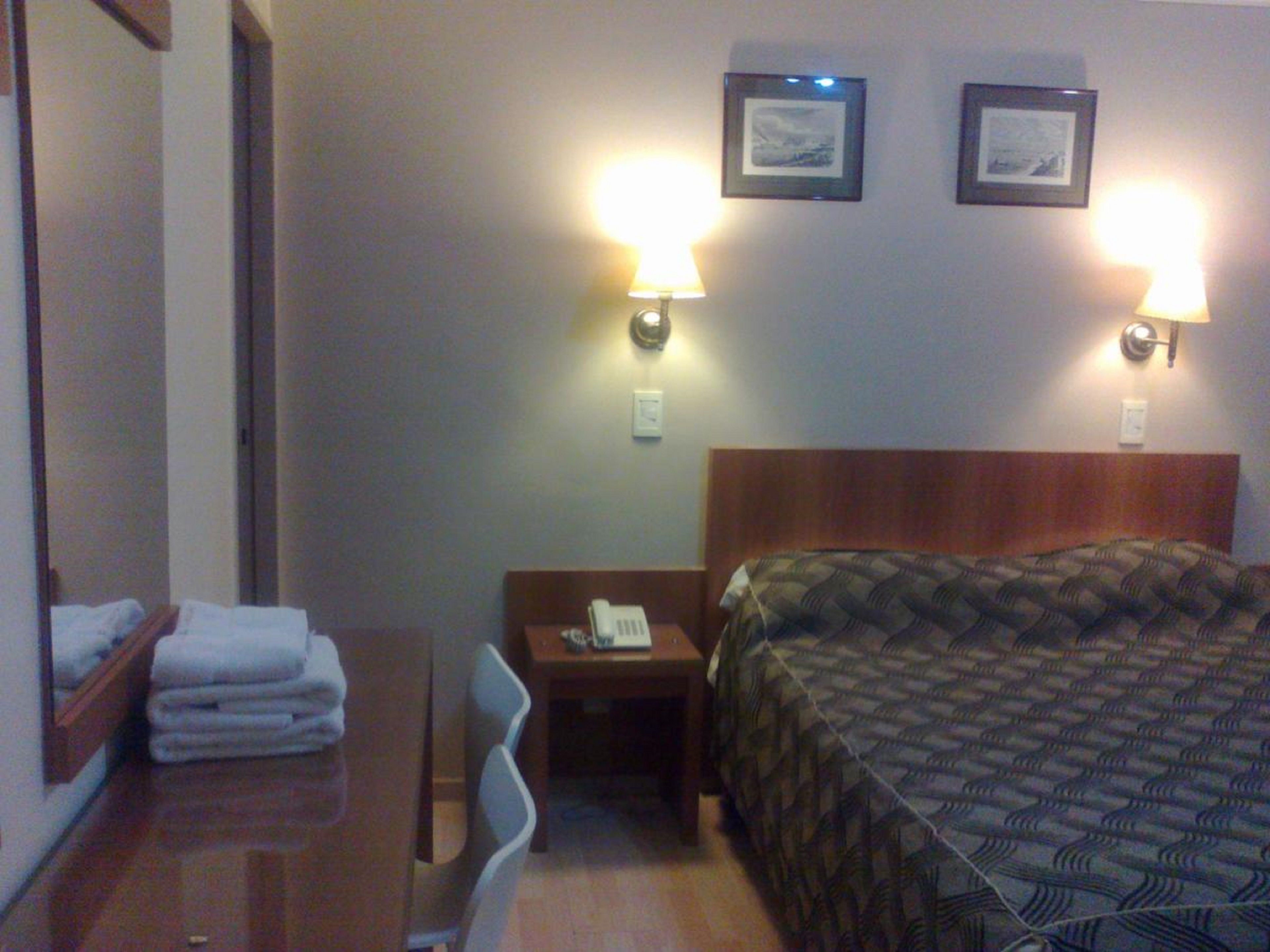 A hotel room with a bed, a desk, a chair, and two wall-mounted lamps.