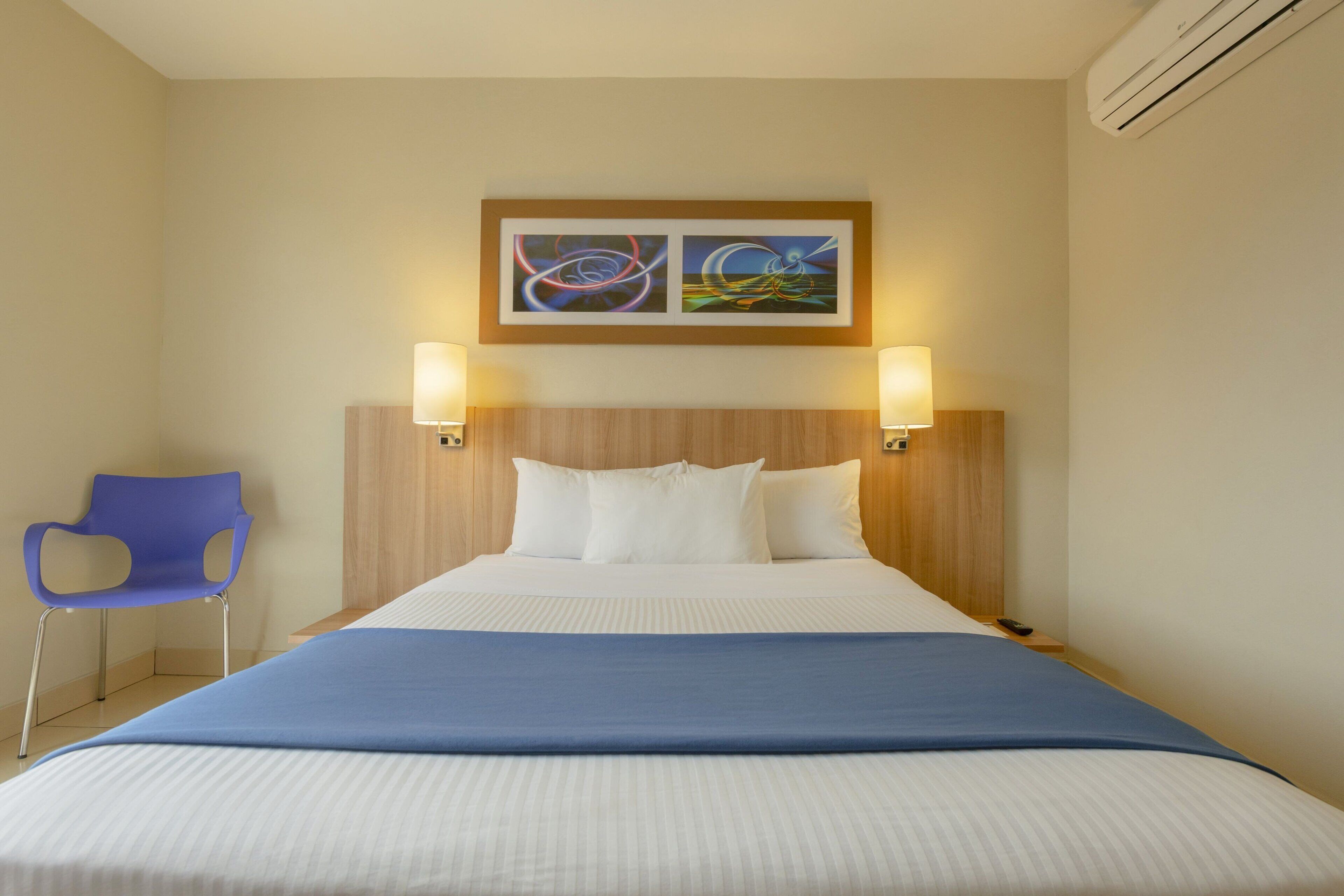 A hotel room with a bed, two wall-mounted lamps, a blue chair, and two framed artworks above the bed.