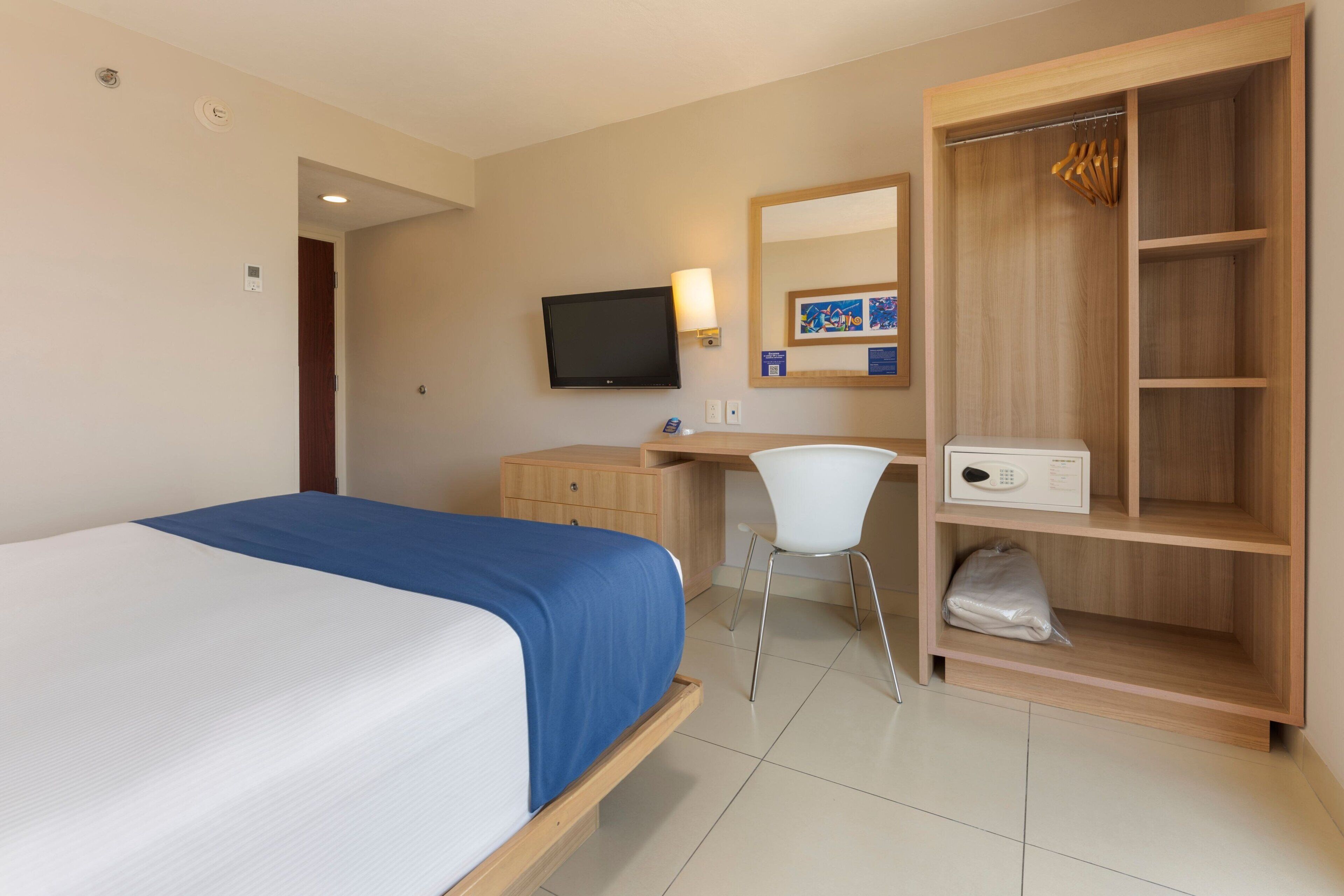 Una habitación de hotel con cama, escritorio, silla, televisor, armario y un mueble bajo.