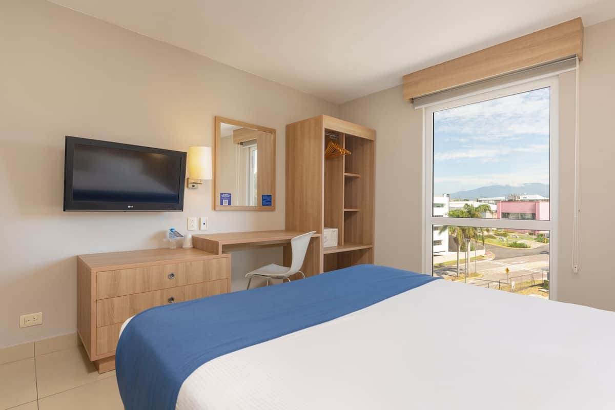 Une chambre d’hôtel équipée d’un lit, d’un bureau, d’une chaise, d’une armoire, d’une télévision et d’une grande fenêtre donnant sur des bâtiments et des montagnes.