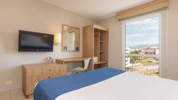 Habitación de hotel con cama, escritorio, silla, armario, televisor y ventana grande con vista a edificios y montañas.