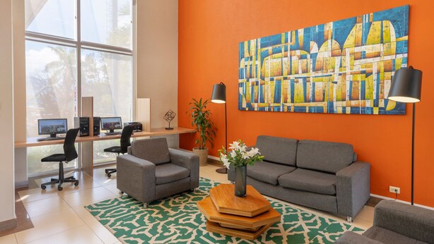 A modern office space with a large window, a desk with computers, two grey sofas, a coffee table, and a large abstract painting on the orange wall. - City Express by Marriott San Jose Costa Rica (Ulloa)