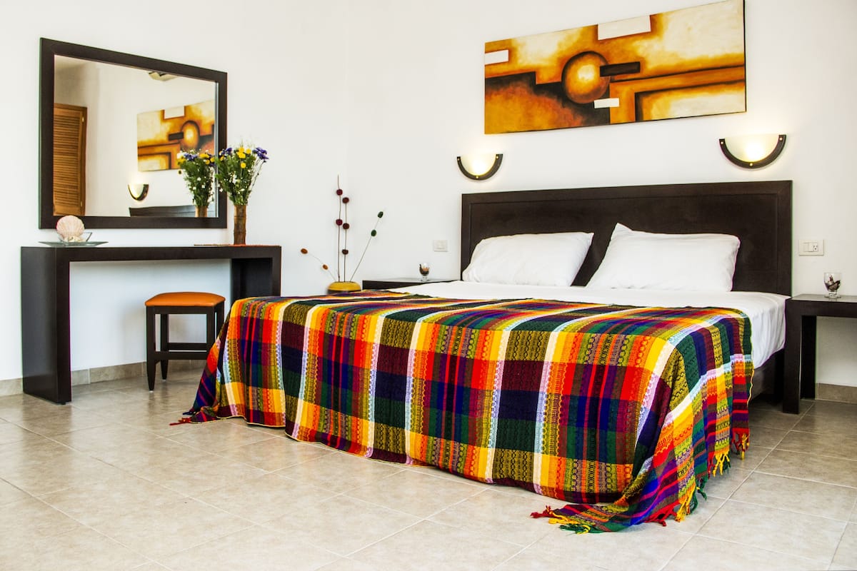 Habitación de hotel con una cama grande, un escritorio con silla, un espejo y dos lámparas de pared.