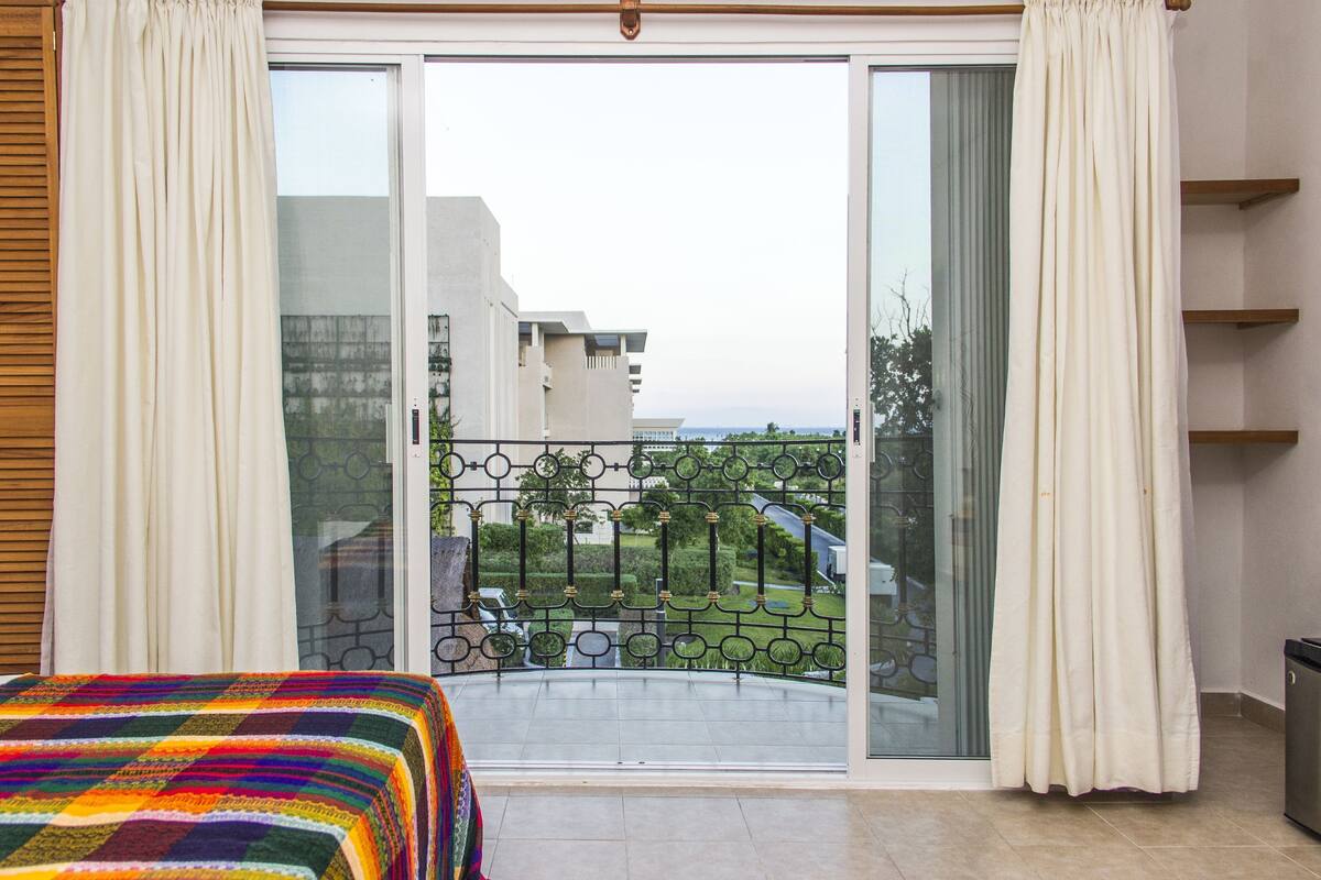 Una habitación con una cama, un ventanal corredero a un balcón y vistas a edificios y vegetación.