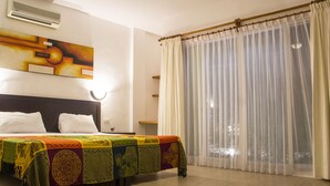 A hotel room with a bed, a colorful bedspread, a large window with sheer curtains, and a wall-mounted air conditioner.