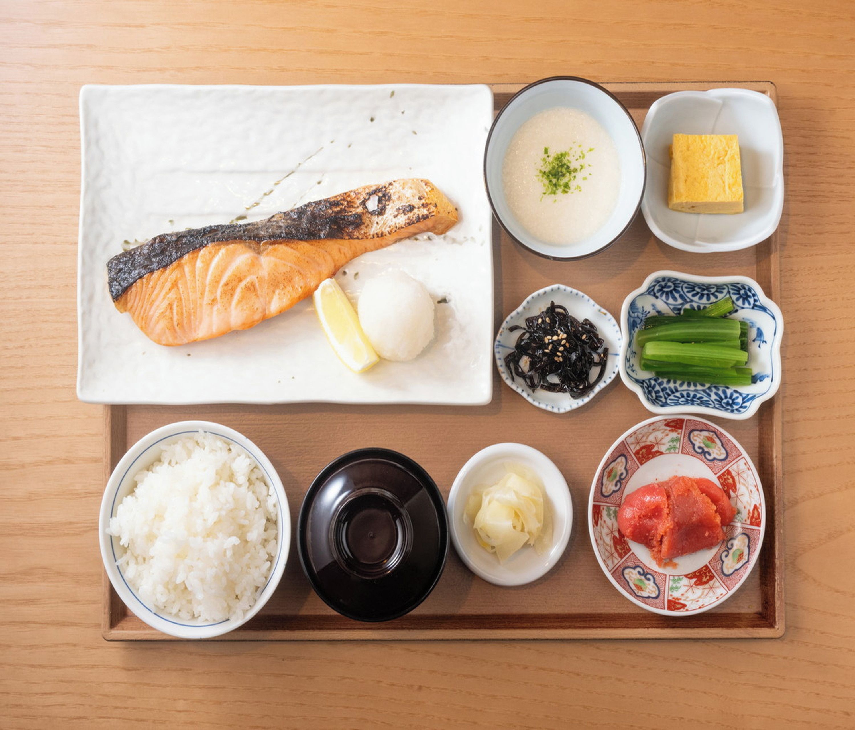 A tray with a salmon fillet, rice, a bowl of soup, a piece of tofu, pickled vegetables, and a small dish of seaweed.
