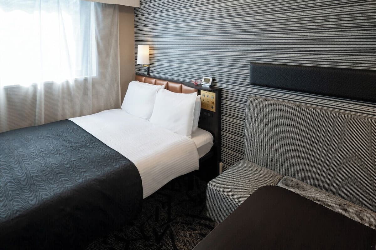 A hotel room with a bed, a bedside table, a lamp, and a view of the outside through a window.