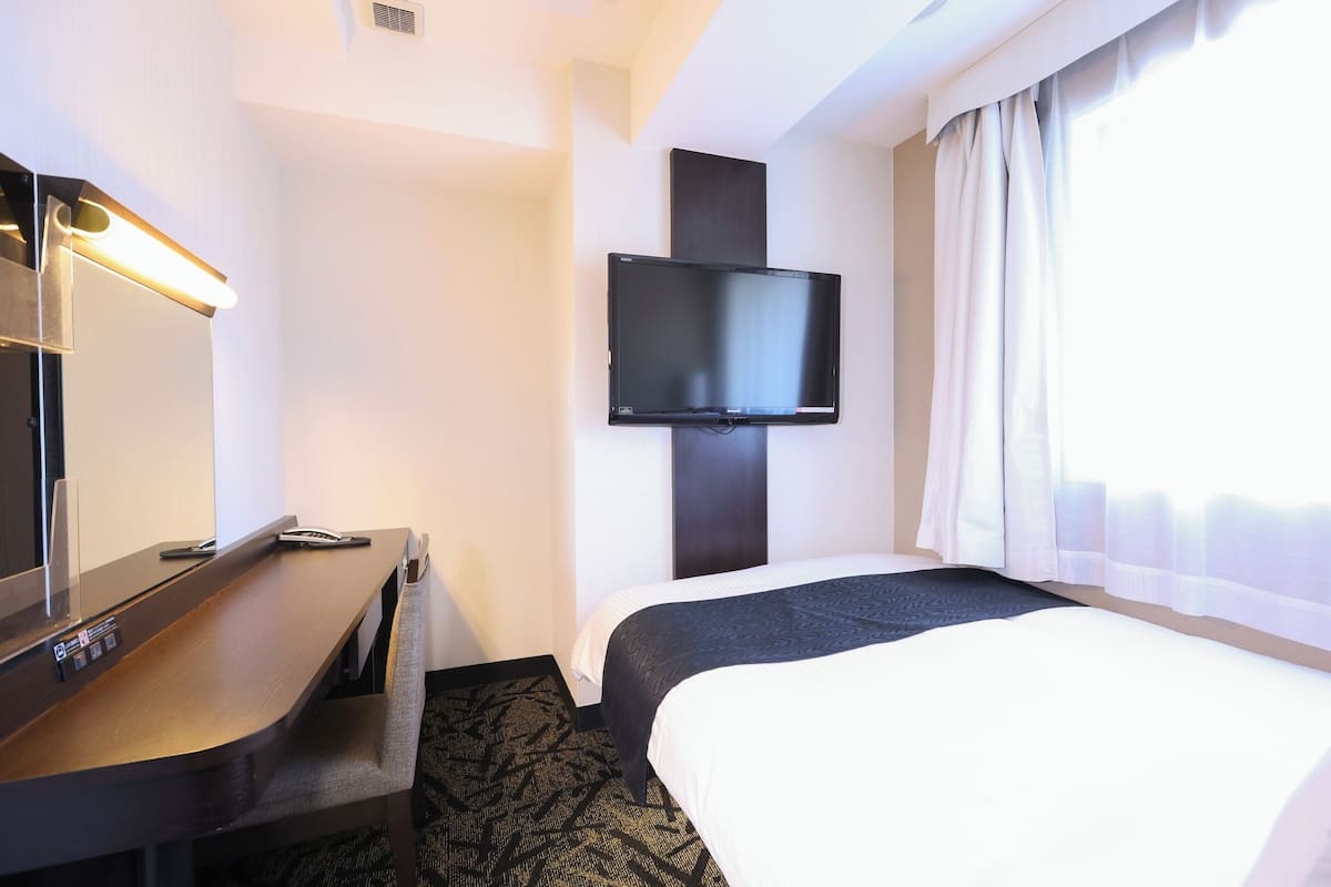 A hotel room with a bed, a desk with a flat-screen TV, and a large window with curtains.