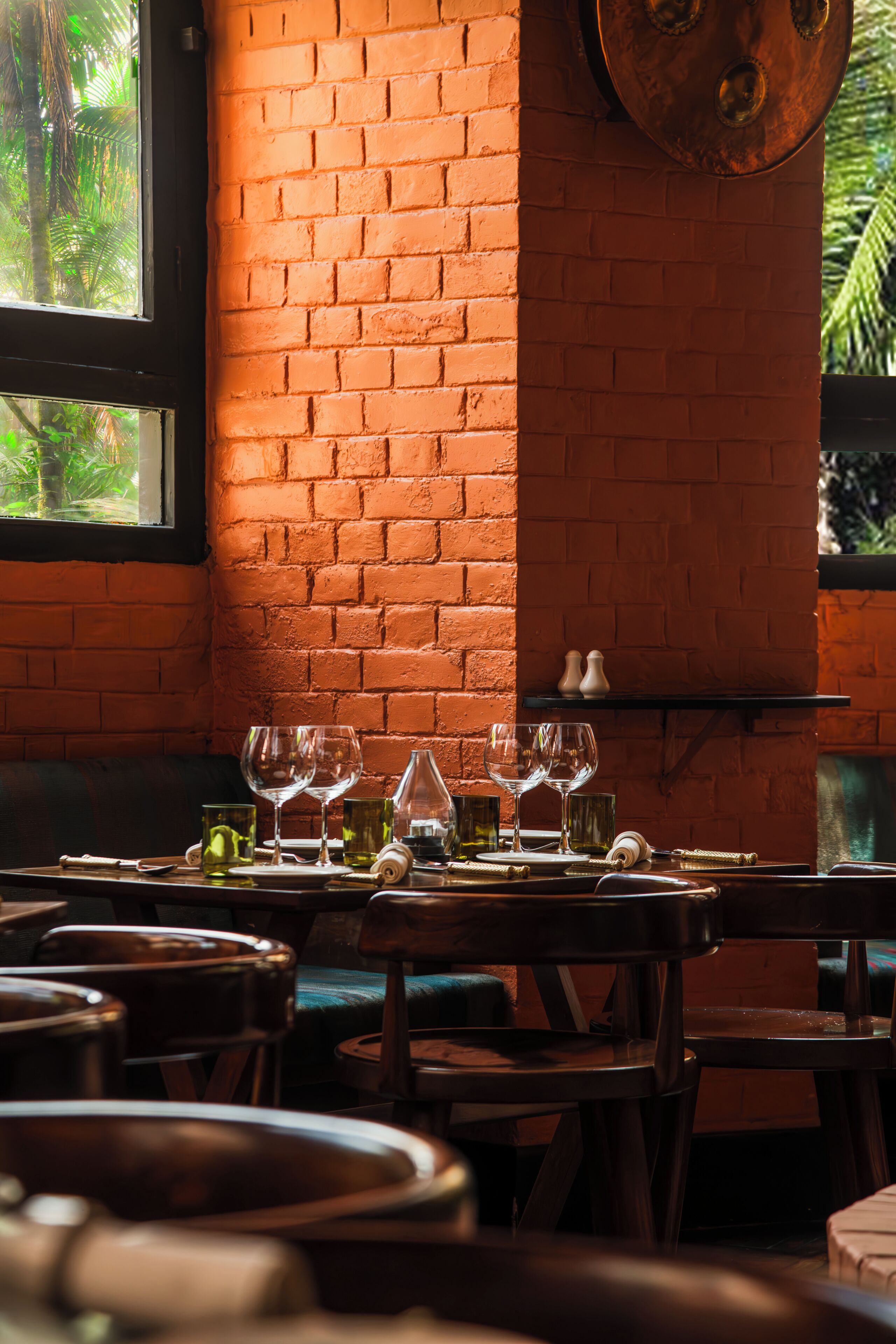 A restaurant interior with brick walls, a window with a view of greenery, and a table set with glasses and cutlery.
