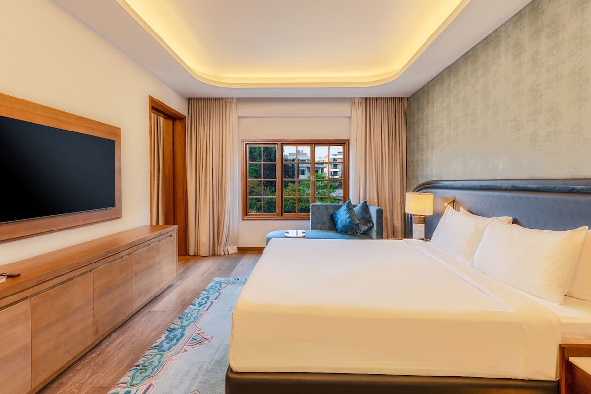 Ein Hotelzimmer mit einem großen Bett, einem an der Wand befestigten Flachbildfernseher, einem Fenster mit Vorhängen und einem Nachttisch mit Lampe.