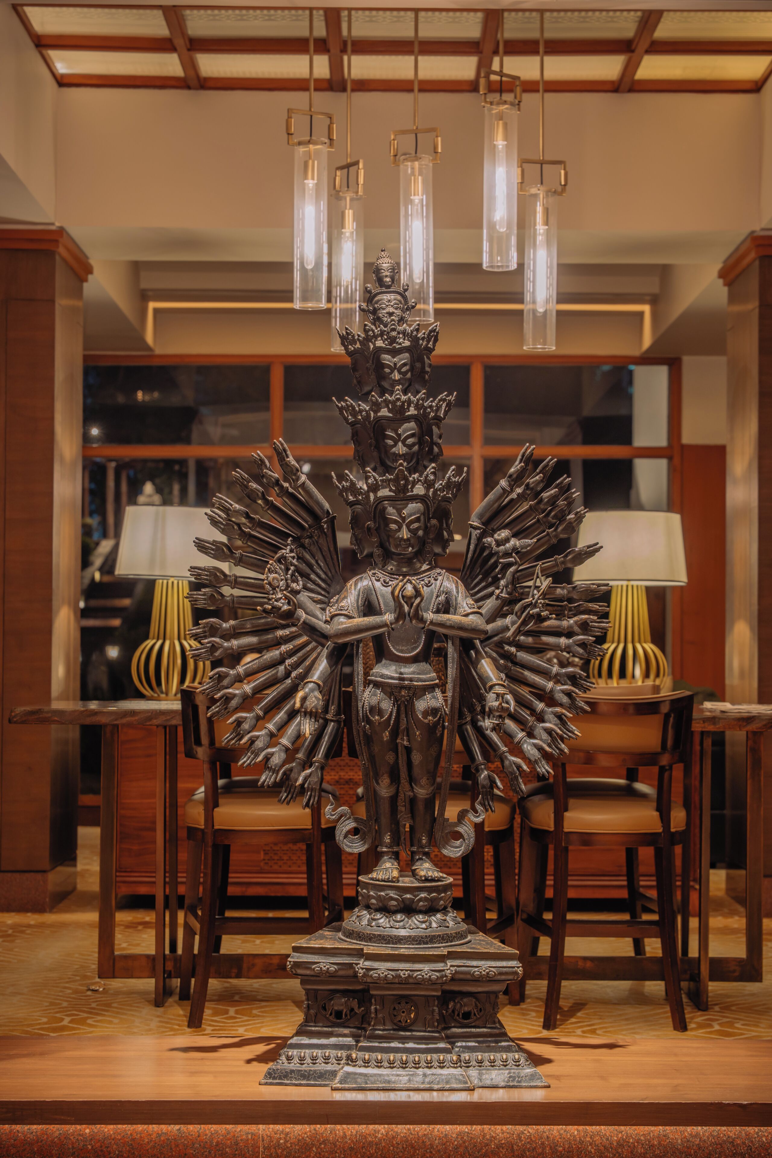 Une statue à plusieurs bras, un piédestal et un lustre orné de suspensions lumineuses, le tout dans une pièce dont le mobilier est en bois et qui possède une fenêtre.