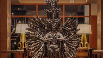 A statue with multiple arms, a pedestal, and a chandelier with hanging lights in a room with wooden furniture and a window.