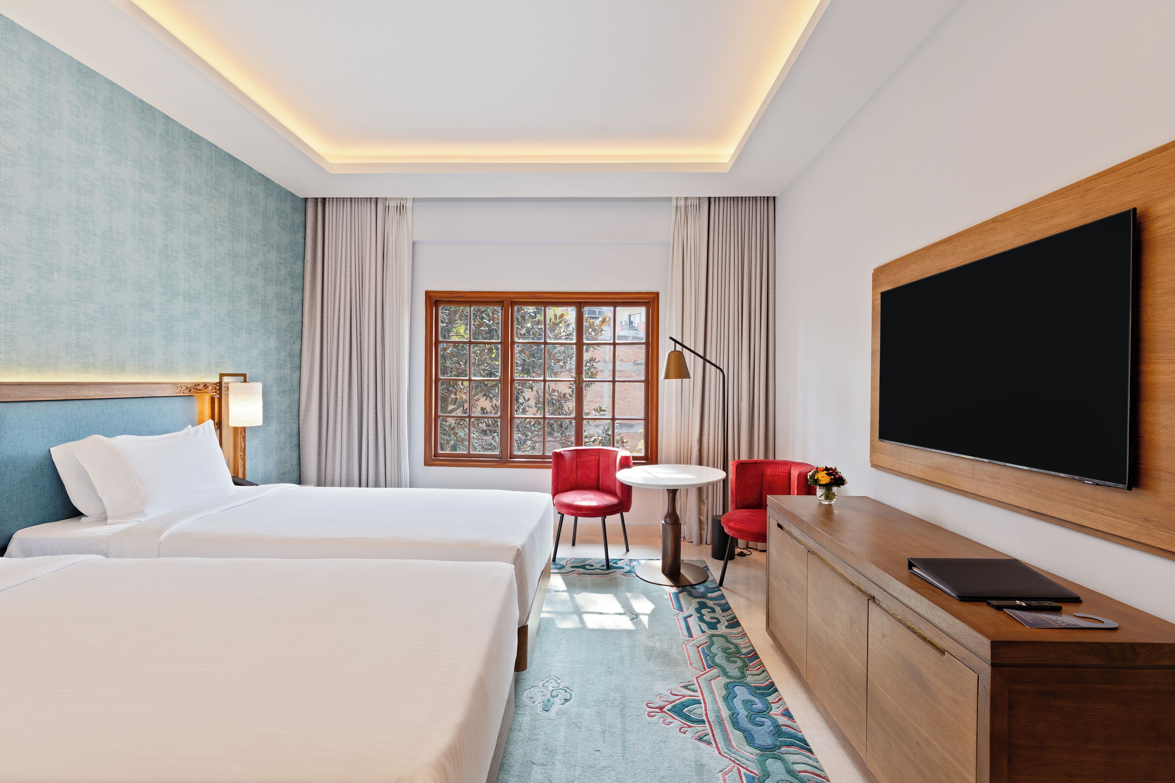 A hotel room with two beds, a TV, a desk with a laptop, a small table, and a window with curtains.
