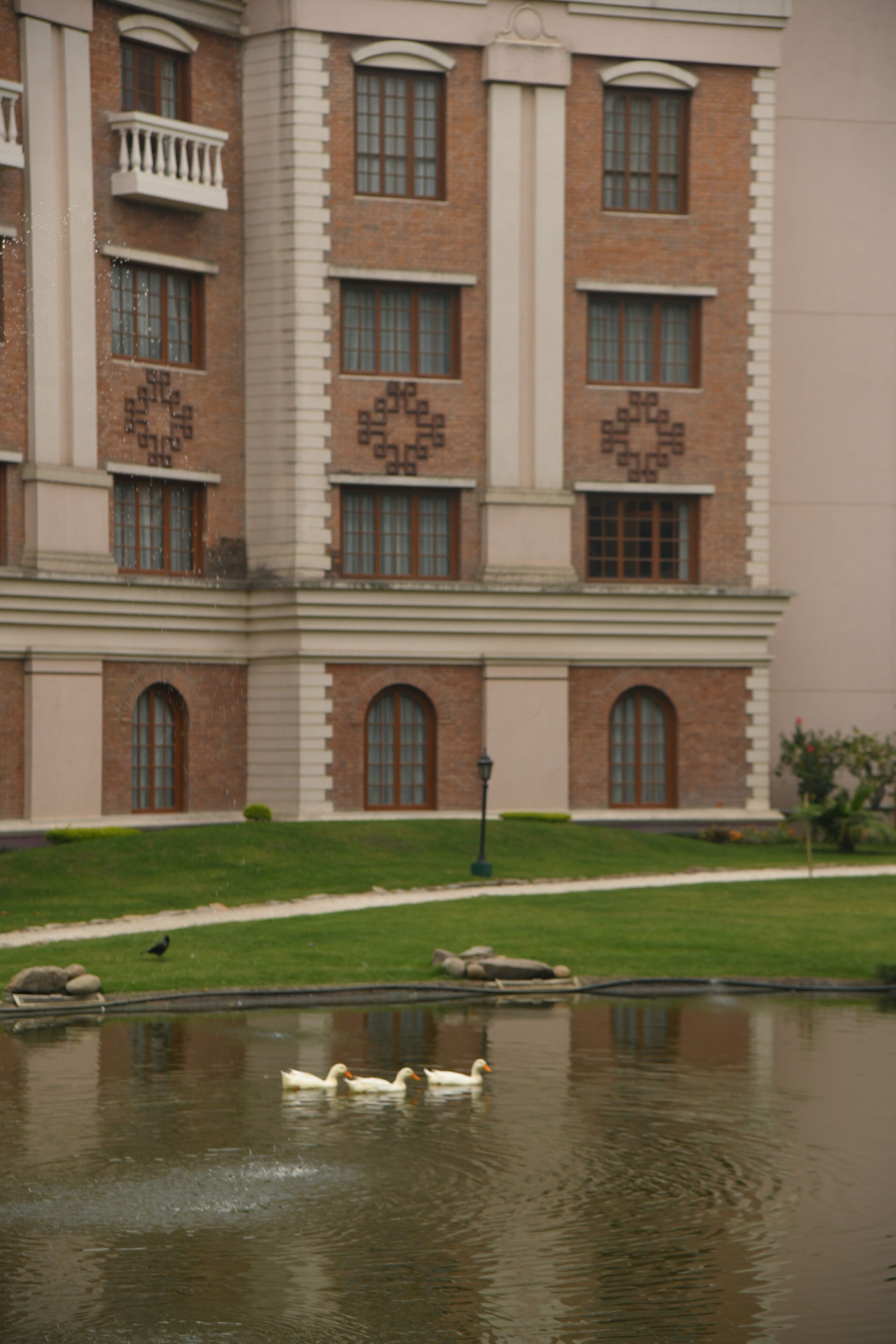 A building with a brick facade, multiple windows, and a balcony. In front, there is a pond with ducks and a bird.