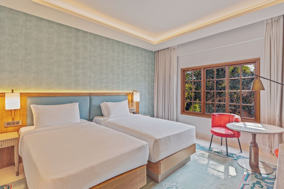 Ein Hotelzimmer mit zwei Betten, einem roten Sessel, einem runden Tisch und einem Fenster mit Blick auf Grünflächen.