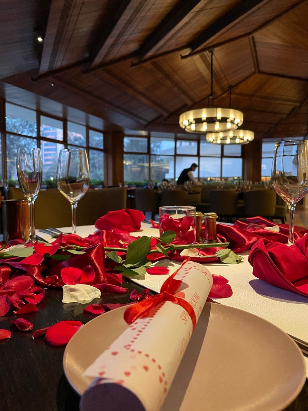 A dining table set with wine glasses, rose petals, and a rolled napkin with a ribbon.